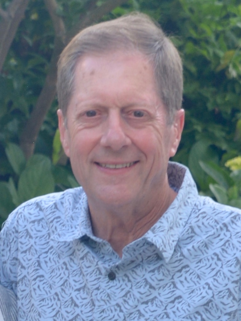 Portrait photo of a smiling older man with grey-brown hair in a patterned open-neck shirt with greenery behind.
