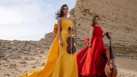 Celtic Connections The Ayoub sisters in the desert, wearing flowing yellow and red dresses. A ruined wall sits behind them.
