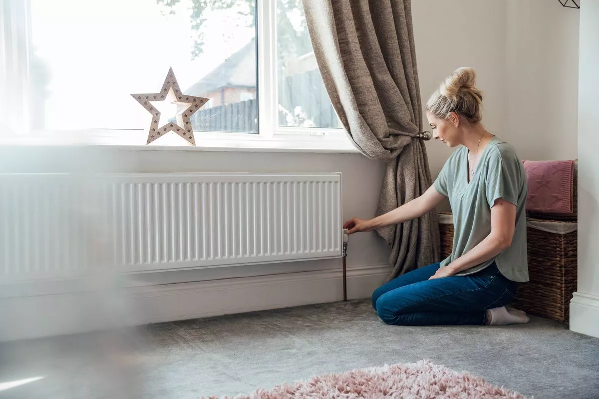 Side view of a young woman kneeling on the living room floor, adjusting the thermostat on the radiator.