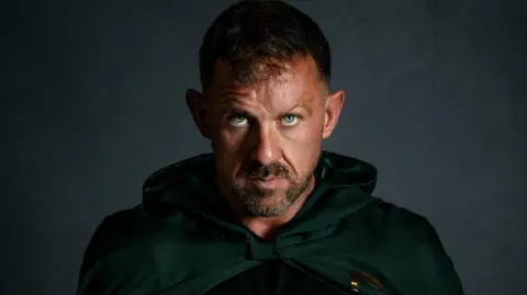 Art of the fairytale Photograph of Andrew Jenkins, pictured wearing a dark green cloak. He has blue eyes, a brown stubble beard and brown hair. 