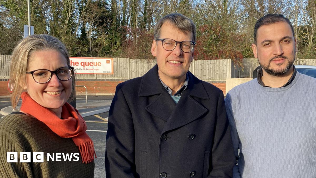 A woman with blonde hair wearing glasses and a green jumper and red scarf smiles towards the camera, she is standing next to a man with grey hair smiling toward the camera. He is wearing glasses and a dark jacket and is standing next to a a bearded man with dark hair who is smiling toward the camera. He is wearing a grey knitted jumper. All three are stood in a line in a car park , wooden fencing and trees can be seen in the background.