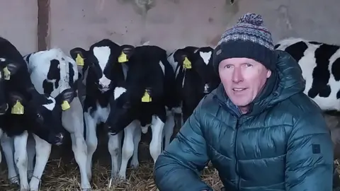 Darren Kidd/BHF A man crouching down in front of a row of black and white cows. The man is wearing a wooly hat and a dark green coat. 