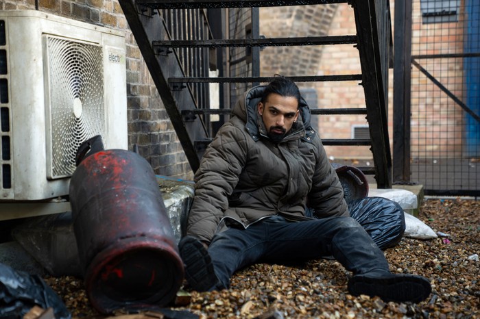 Ravi sits clumsily among some leaves in a wasteland in EastEnders