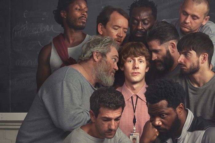 Francis Lovehall as Dris, Alex Ferns as Keith, Josef Altin as Greg, Steven Meo as Macca, Josh Finan as Dan, Sule Rimi as Samson, Ric Renton as Wallace, Tom Moutchi as Junior, Charlie Rix as Zach and Nima Teleghani as Malik in Waiting for the Out. Dan is wearing a pink shirt with a lanyard on and the others are all wearing grey and gathered around him, looking at him intently.