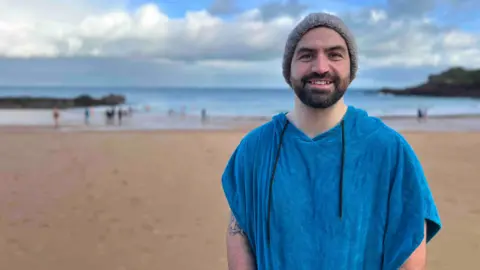 Michael Ahier is wearing a hat and towel while standing on the beach.