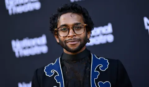 Getty Images Hiran Abeysekera stands in front of a National Theatre sign. H ei swearing black with a blue pattern and glasses