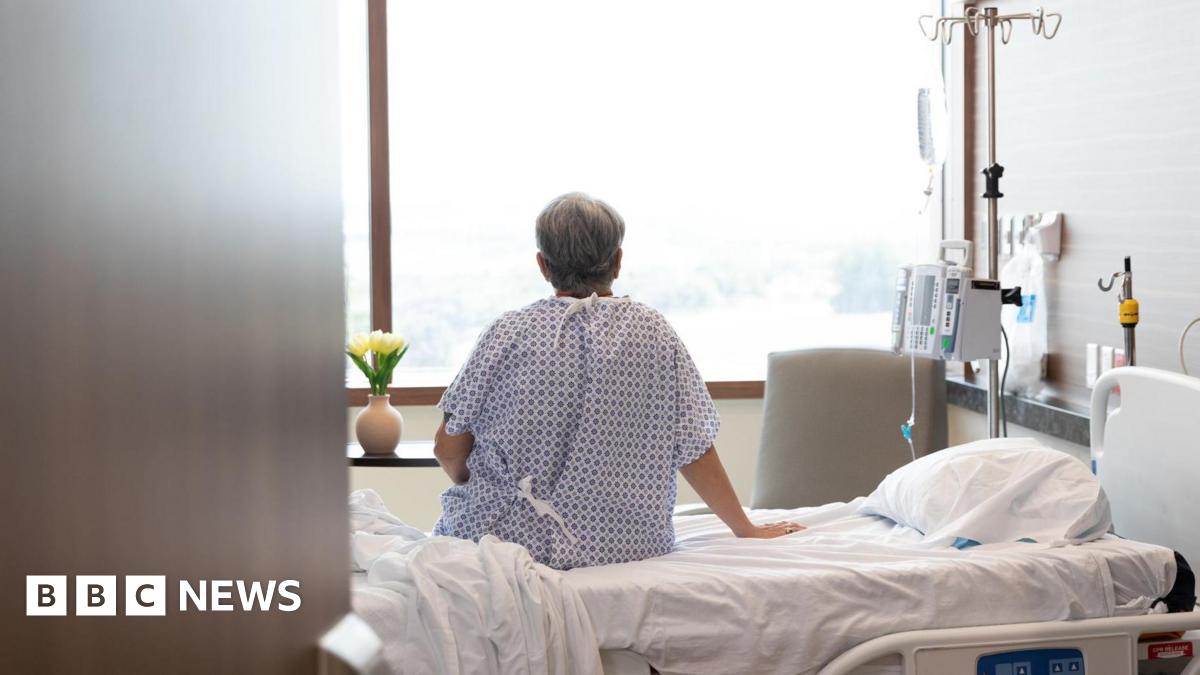 A man sits on a hospital bed facing a window. His back is facing the camera.