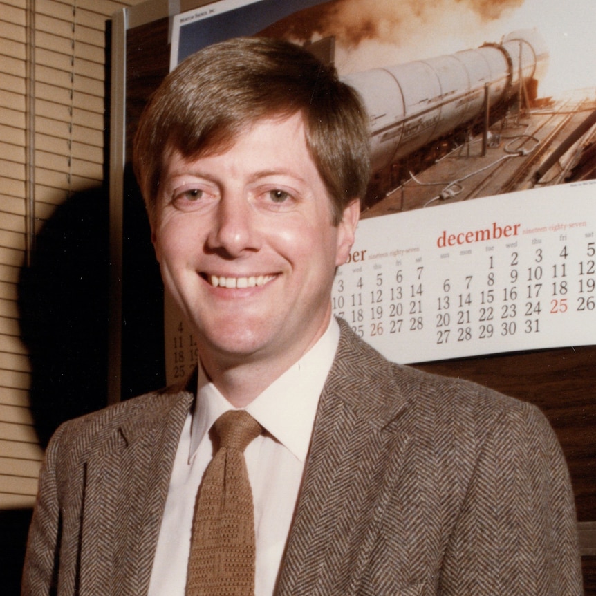 A young man with a brown jacket, brown tie, brown hair and brown eyes smiles at the camera with a rocket calendar behind him.