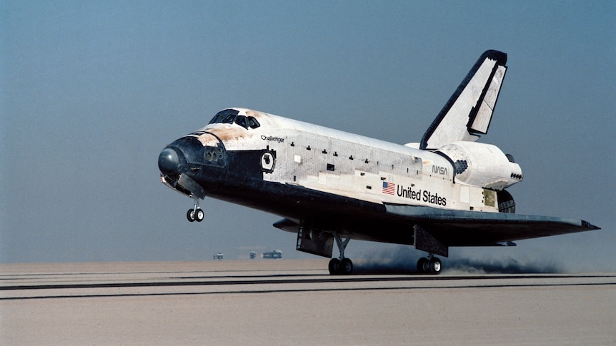 Photo of a space shuttle at the moment its rear wheels touch a desert runway, nose still pointing slightly upward.