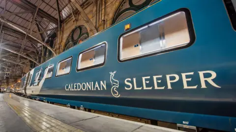 Caledonian Sleeper/PA Wire A dark blue train carriage is photographed at a train station. The carriage has white patterns across it and "Caledonian Sleeper" written on the side in white writing. 