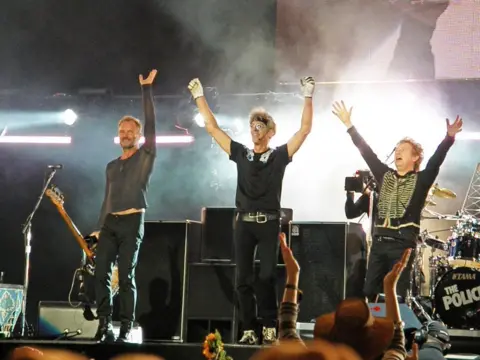 Getty Images The Police on stage in Hyde Park in 2008 - they all have arms in the air and are smiling as they acknowledge the crowd - Sting is holding a guitar and there is a drum kit and speakers behind them 