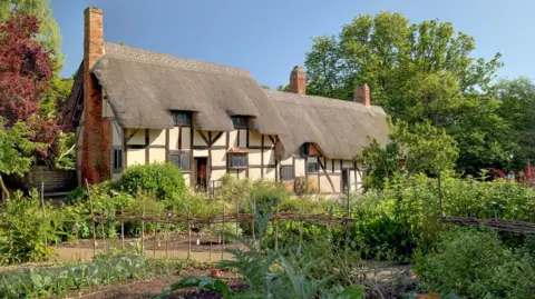Shakespeare Birthplace Trust A black and white house with a green garden full of plants, trees and bushes. The house has lattice windows and a thatched roof