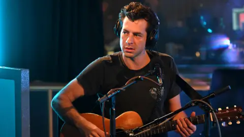Mark Ronson, a man with dark hair, sitting in a recording studio while holding an acoustic guitar,