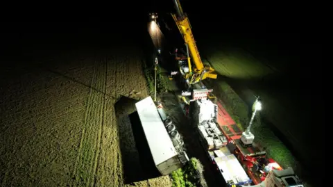 McLaughlin's Light and Heavy Recovery Services A drone shot of the crash scene at night. A large crane is helping to lift a large white lorry out of a field. There are a few large lights helping to light up the scene.