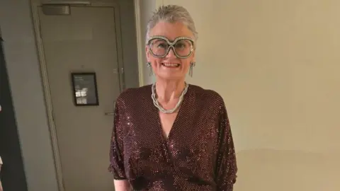 Carolyn Harris MP Carolyn Harris wearing a sparkling sequin maroon dress. She is standing  in what appears to be a hotel bedroom in front of the door and has on oversized, sparkly glasses. Carolyn is wearing a rope-style necklace and dangly earrings