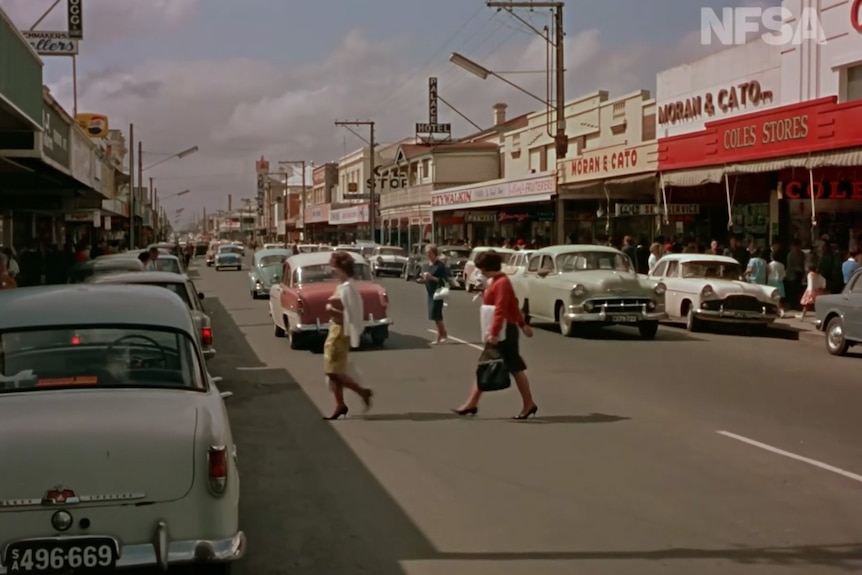 An old restored photo of a street with many colourful shops and old cars parked on each side of the road and people crossing.