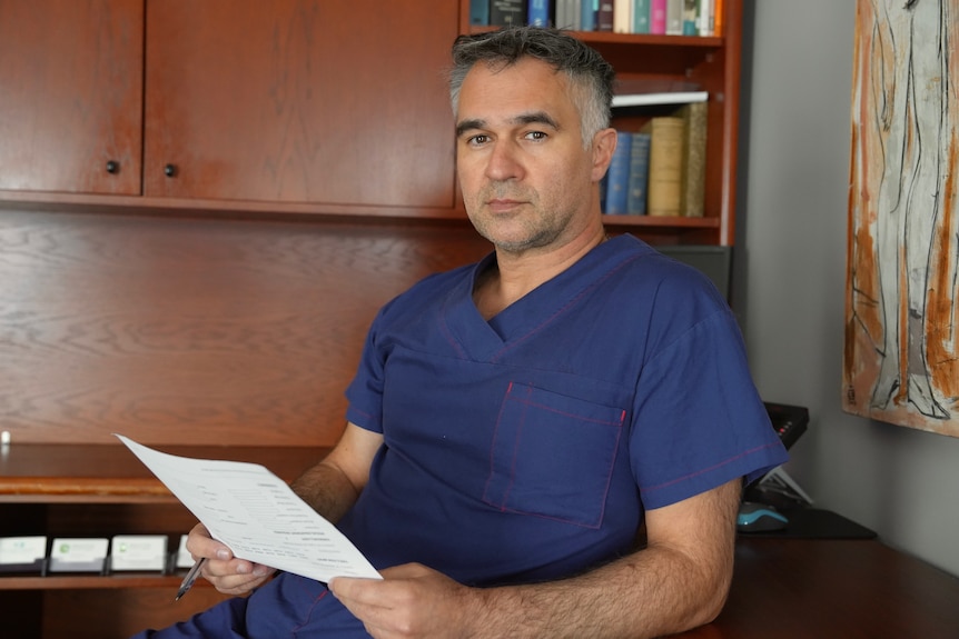 A photo showing a man blue hospital scrubs looking at the camera.