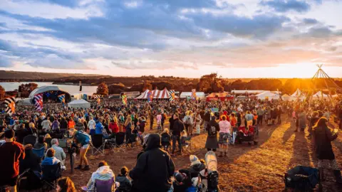 Ania Shrimpton Crowds of people on a festival site, with the sun setting nearby