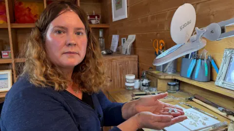 BBC A woman with brown curly hair wearing a blue jumper. She is sitting in a wooden workshop surrounded by various tools. She is holding a pair of silver earrings which are rectangular in shape.