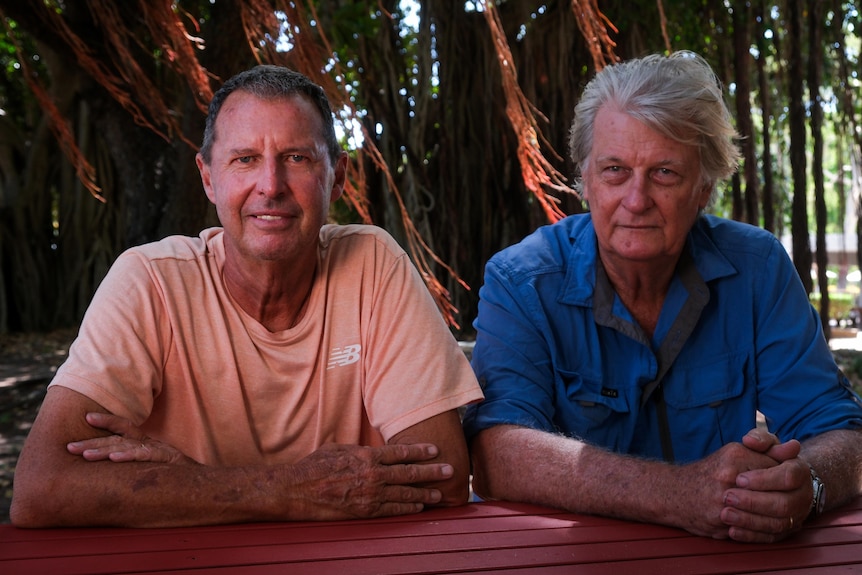 Two men sit at a park bench, facing the camera