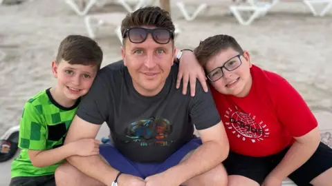 Tim Sadler Image of two boys and a man sitting together cross-legged on a beach.  One boy is wearing a green t-shirt, the man is wearing a black t-shirt and blue shorts and sunglasses on his head. The second boy is wearing a red t-shirt and is wearing glasses.  All three are smiling at the camera.