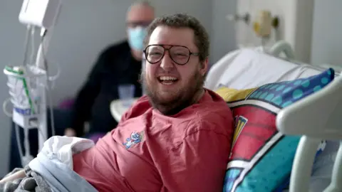 BBC A man with brown hair and a beard smiling wearing aviator-style glasses sits in a hospital bed. He is wearing a red Lilo and Stitch top