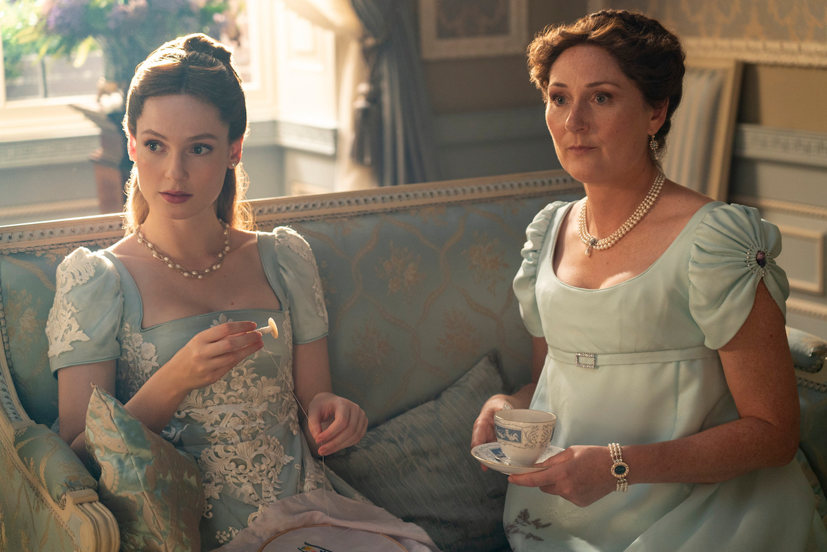 Two women in elegant historical dresses sit on a decorated sofa in a luxurious, sunlit parlor, holding a teacup and embroidery, with light streaming through the window, creating a refined, vintage atmosphere.