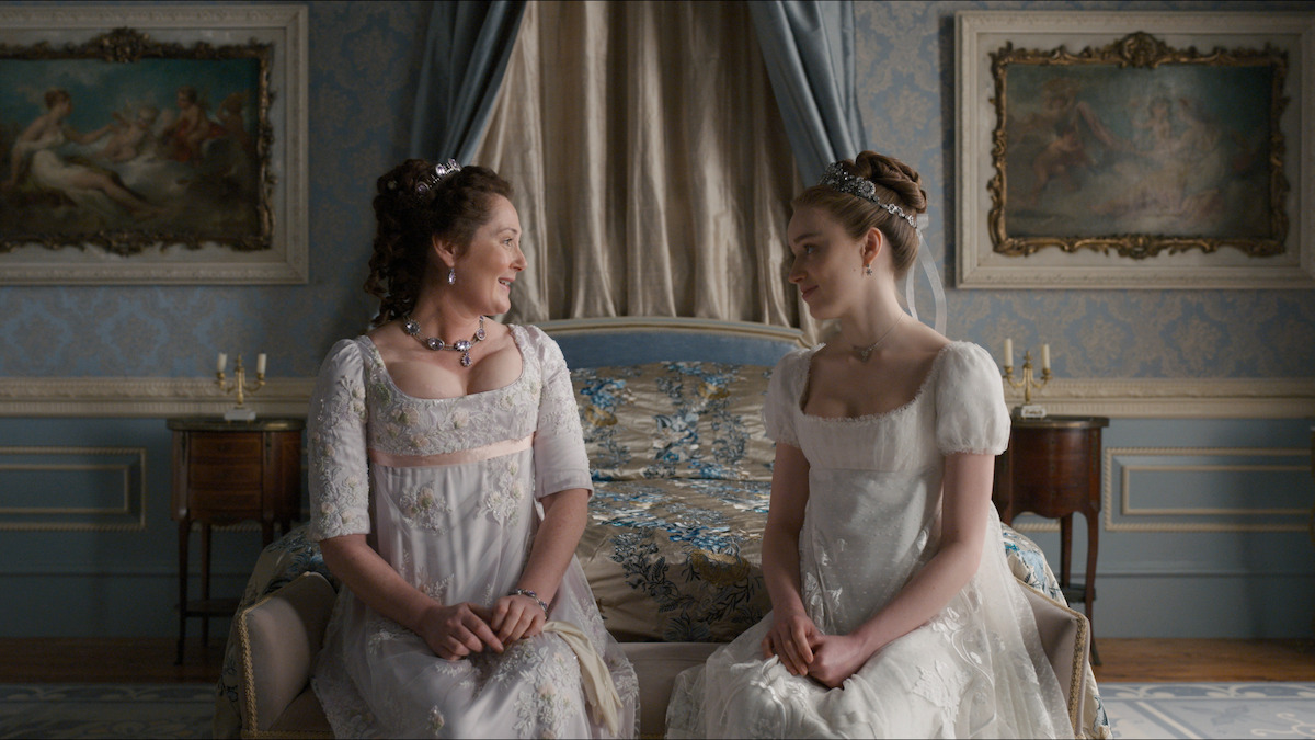 Two women in Regency-era dresses sit on a bed in an ornate, pastel blue room with classical paintings, elegant furniture, and luxurious drapes, having a warm conversation.