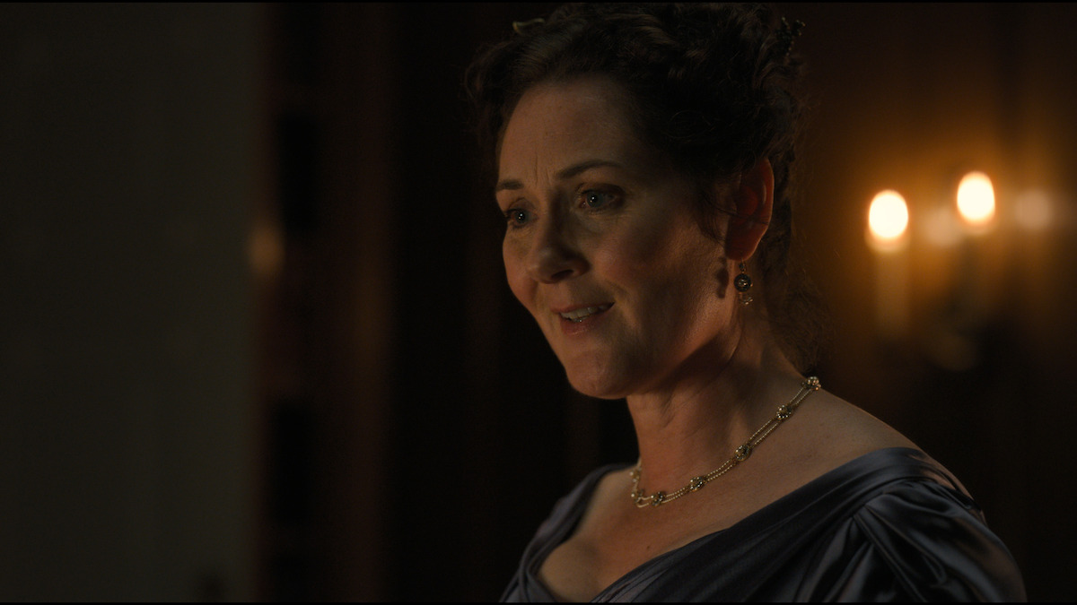 A woman in period dress stands indoors in soft, warm light, smiling gently. She wears elegant jewelry and a blue gown. Candlelight and blurred architecture form the background, suggesting a historical or formal setting.