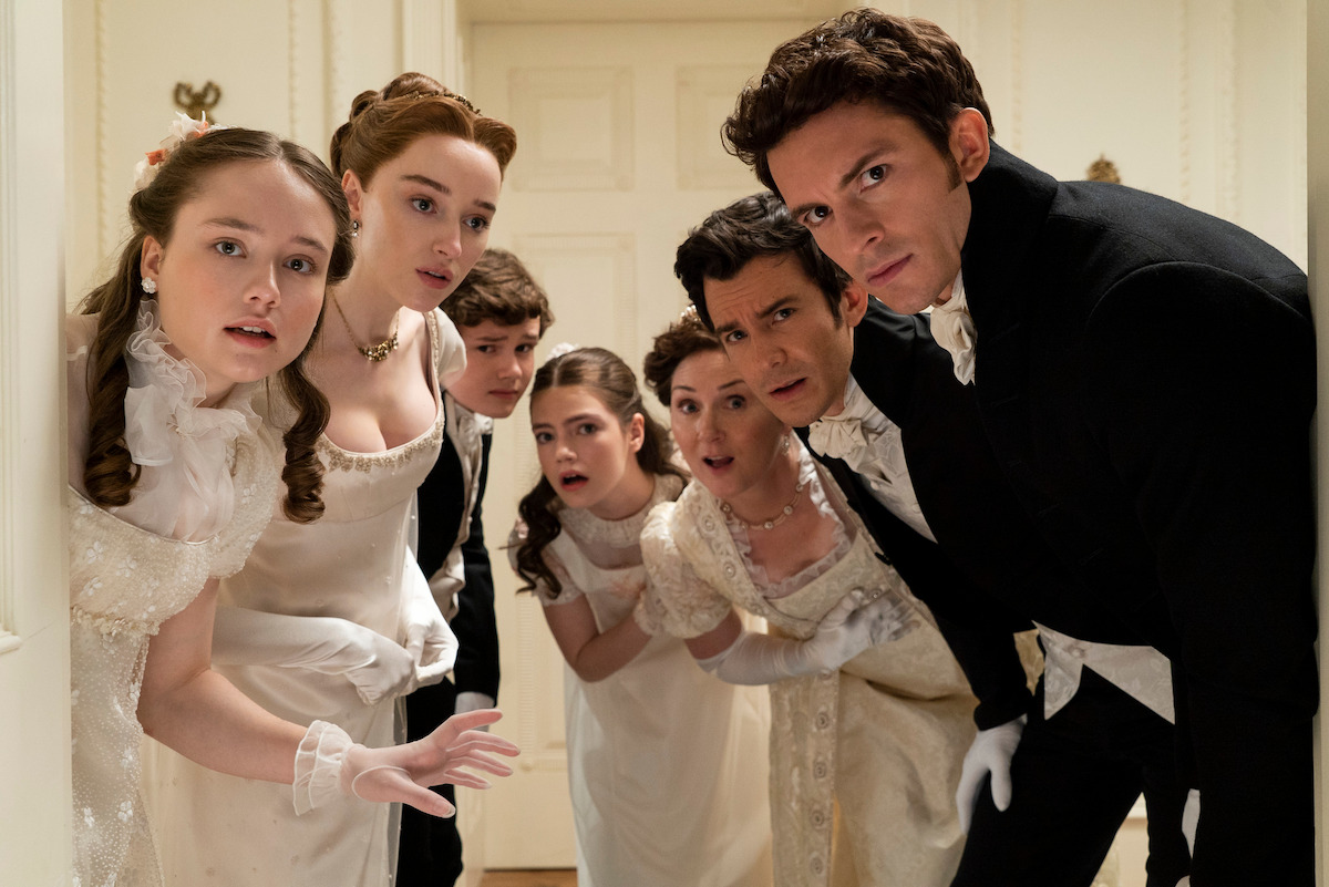 Seven people in elegant Regency-era clothing huddle in a hallway, looking curiously toward the camera, suggesting a dramatic or comedic moment in a period drama setting.