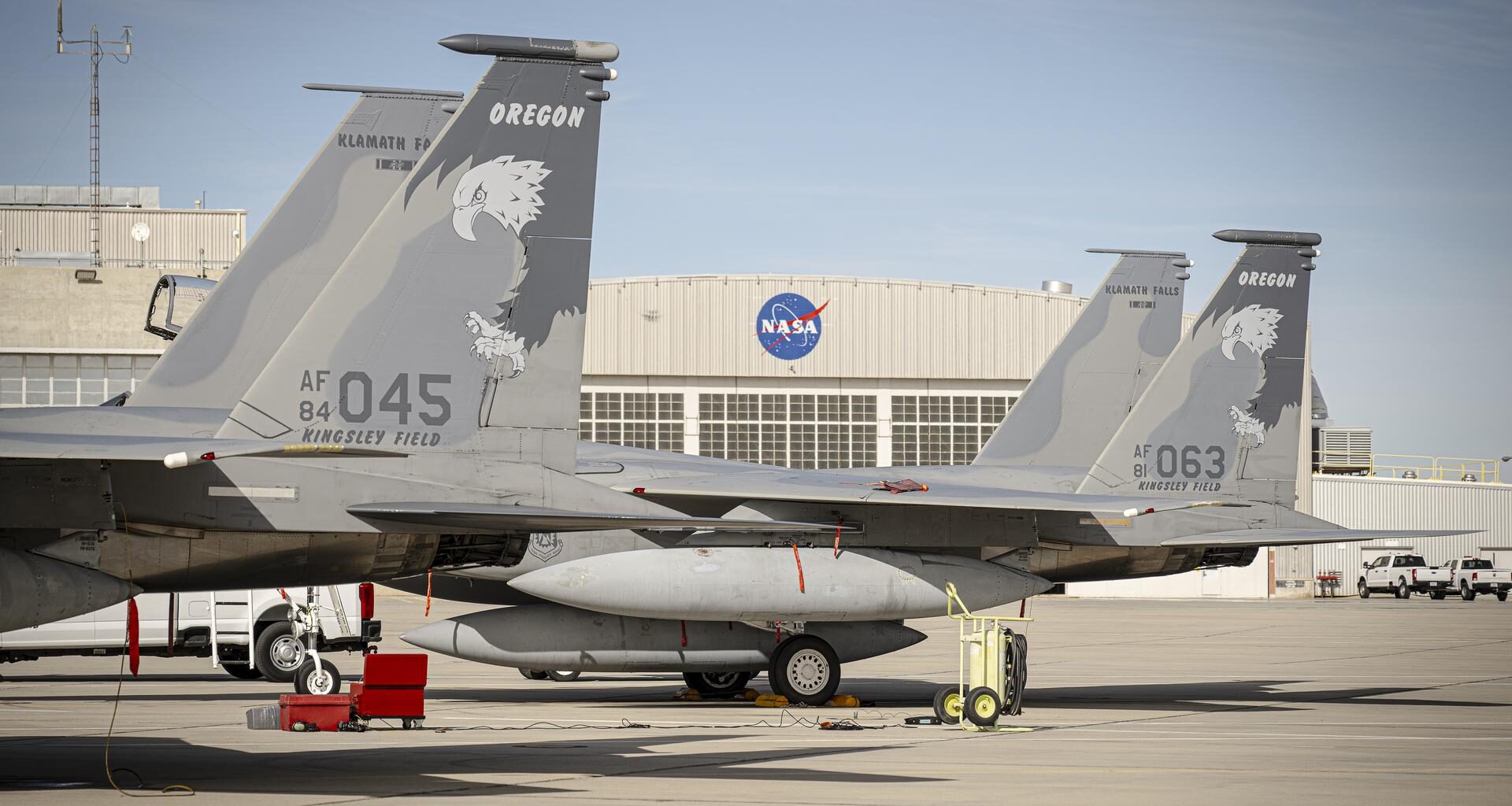 Two F-15 aircraft sit side by side on the ramp at NASA’s Armstrong Flight Research Center, displaying tail numbers 045 and 063 and tail markings that read “Oregon” above an eagle graphic. A NASA hangar with the agency’s logo is visible in the background