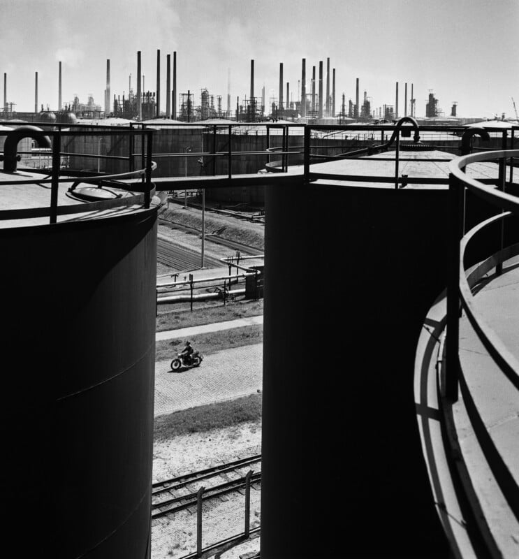 A lone motorcyclist rides on a road between large industrial tanks, with a sprawling refinery and smokestacks visible in the background under a hazy sky.