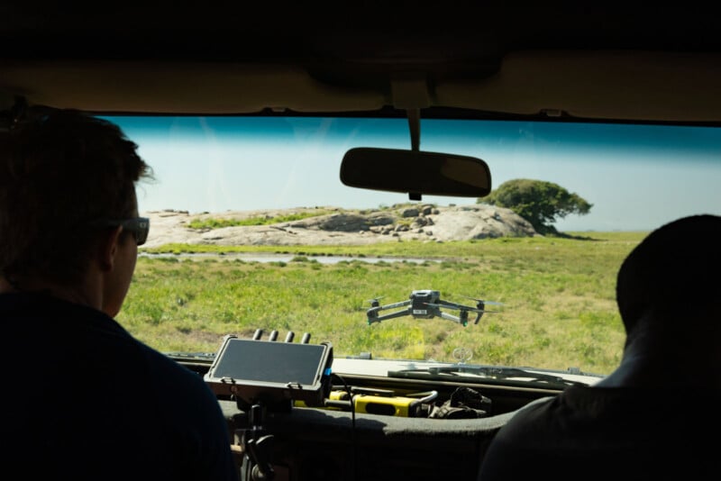 Two people inside a vehicle watch a drone flying outside over grassy terrain, with a rocky hill and a tree visible in the distance through the windshield. A monitor and other equipment are mounted inside the vehicle.