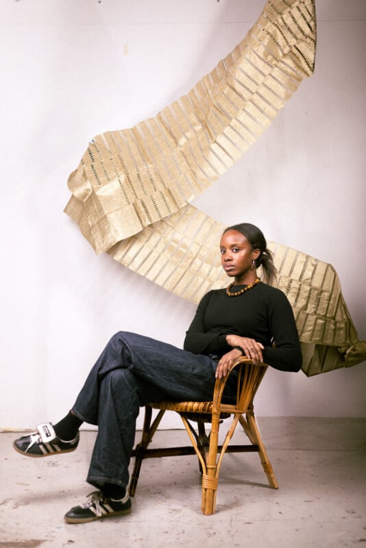 A woman sits in a wicker chair against a white wall, with a large, textured, gold fabric sculpture hanging behind her. She wears a black top, dark jeans, sneakers, and a beaded necklace, looking calmly at the camera.