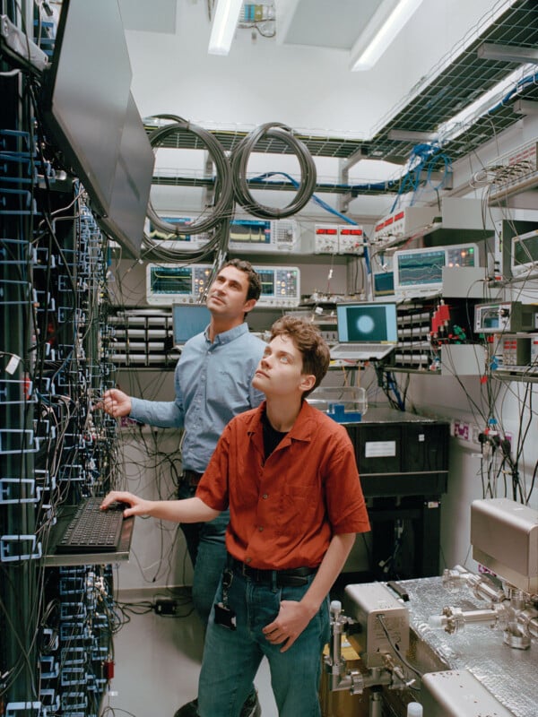 Two people stand in a cluttered lab surrounded by servers, cables, and electronic equipment. One types on a keyboard while the other looks up at a monitor. Both focus intently on their work.