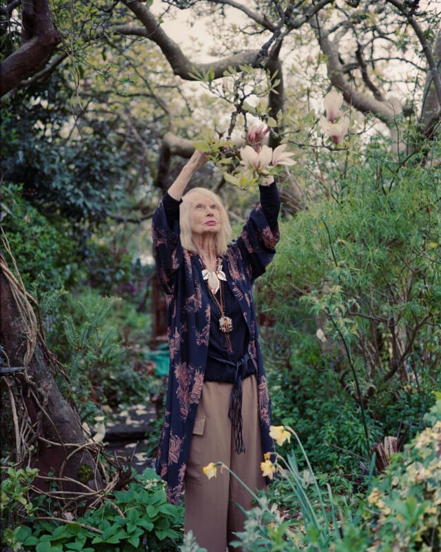 An older woman with long blonde hair stands in a lush garden, wearing a floral kimono and wide-legged pants, reaching up to touch blooming magnolia flowers on a tree branch surrounded by greenery.