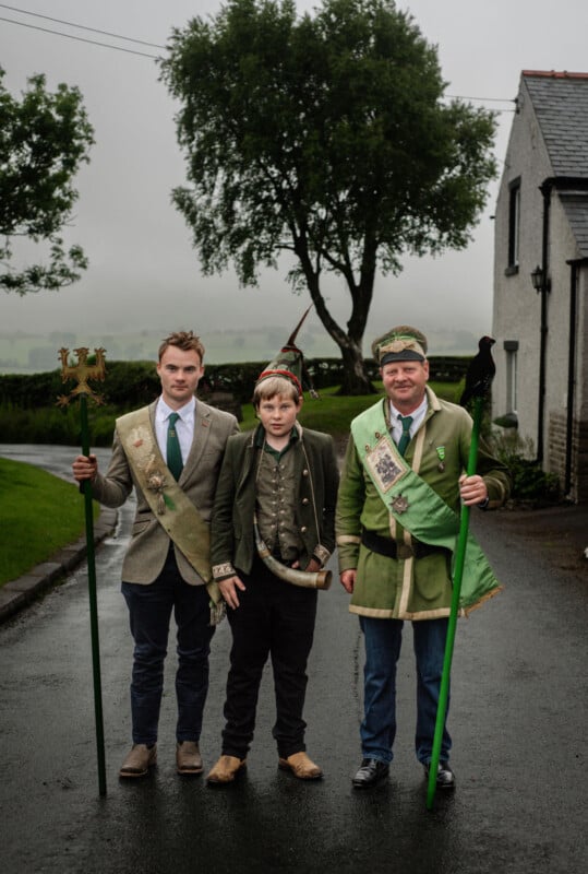 Three people stand outdoors on a wet road, dressed in traditional attire with sashes and hats. Two hold tall green staffs. There is a tree and a white house in the background, with cloudy, misty weather.