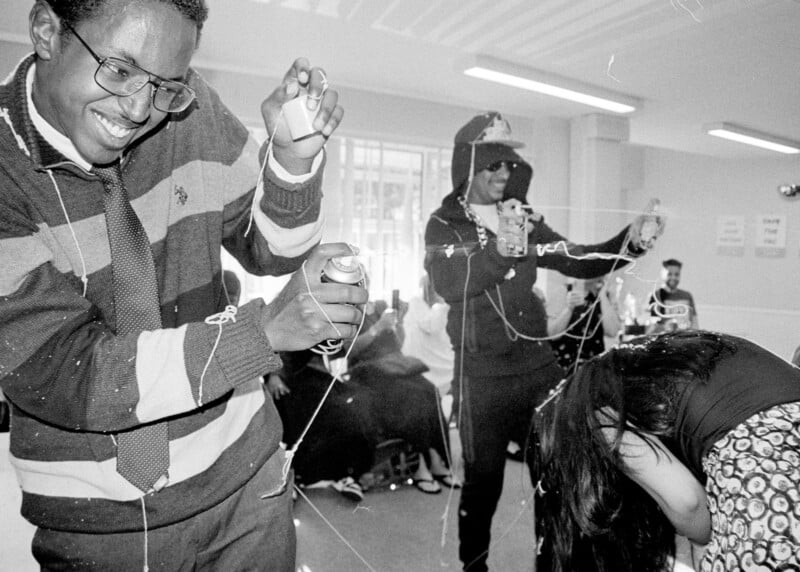 Three people in a lively indoor setting spray silly string and laugh, while others sit watching in the background. One person bends forward, a man in glasses smiles, and another wears sunglasses and a cap.