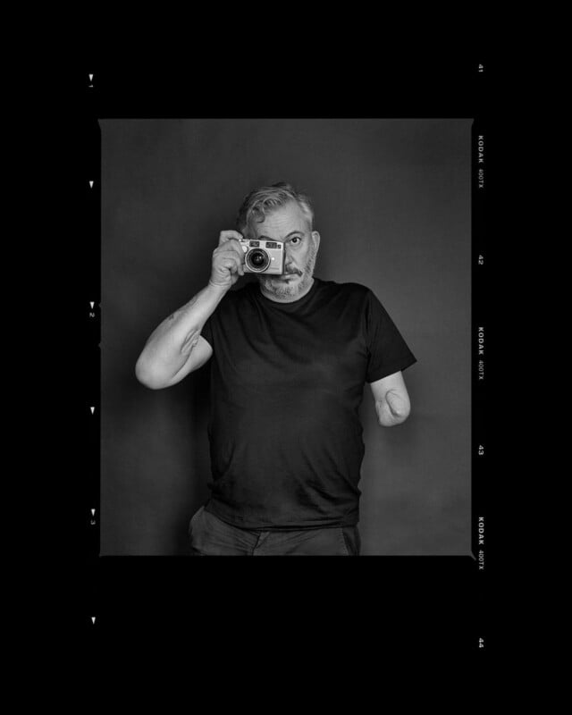 A man with a beard and a partial right arm wears a black t-shirt and poses against a dark background, holding a camera up to his eye as if taking a photo. The image is framed to resemble a film contact sheet.