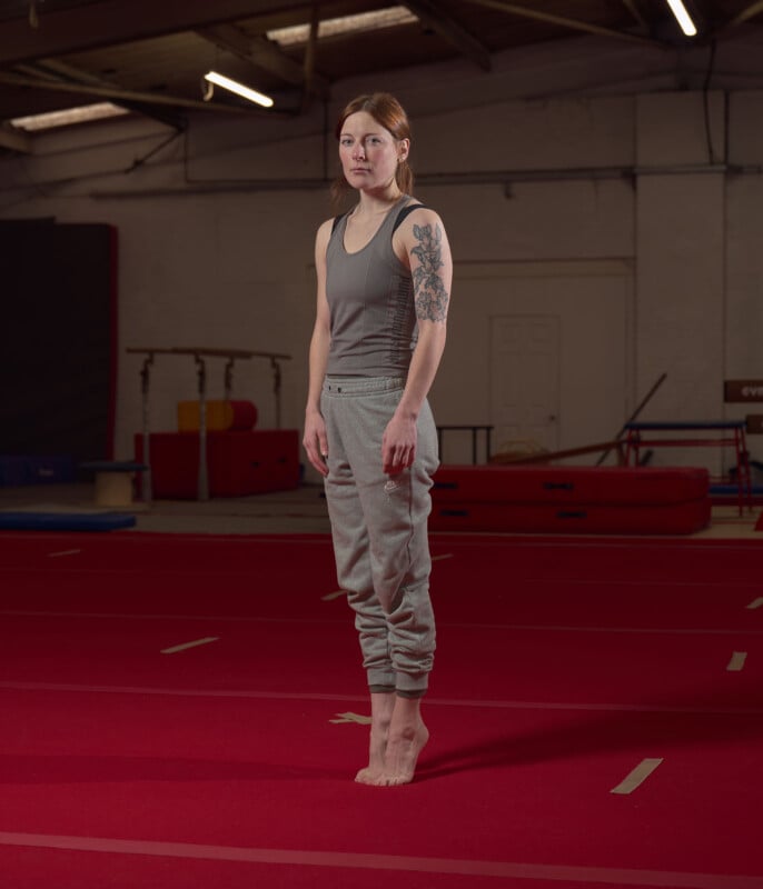 A woman standing barefoot on red gymnastics mats in a gym, wearing a gray tank top and sweatpants, with a tattoo on her left upper arm. She looks forward with a neutral expression. Gymnastics equipment is visible in the background.