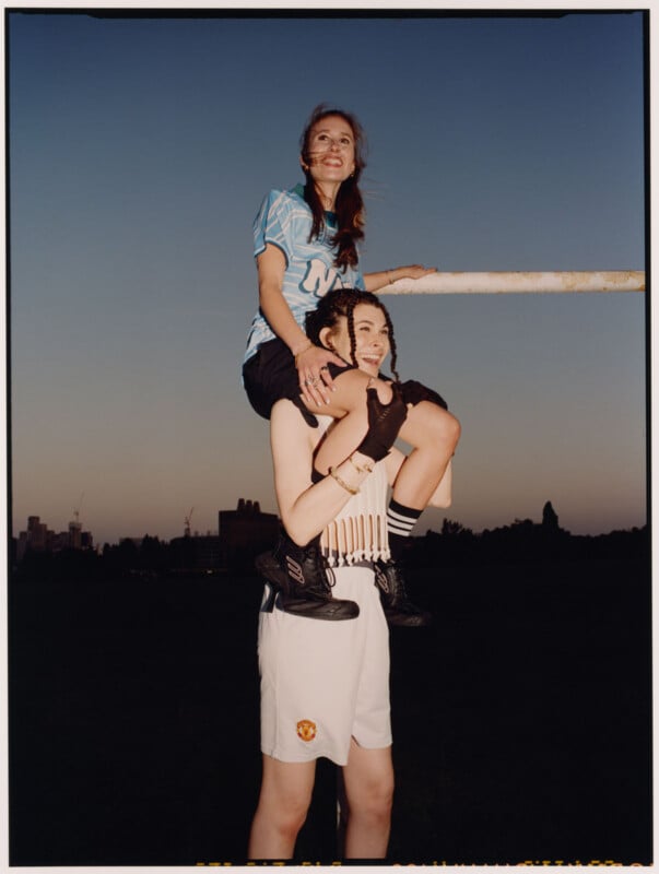 Two young women pose outdoors at dusk; one sits on the other’s shoulders, smiling. The woman on top wears a blue sports jersey, black shorts, and painted cheeks; the one below wears white shorts, black boots, and a sleeveless top.