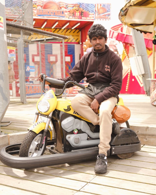 A man with curly hair sits on a yellow toy motorcycle ride at an amusement park, looking at the camera. Colorful fairground attractions and games are visible in the background.