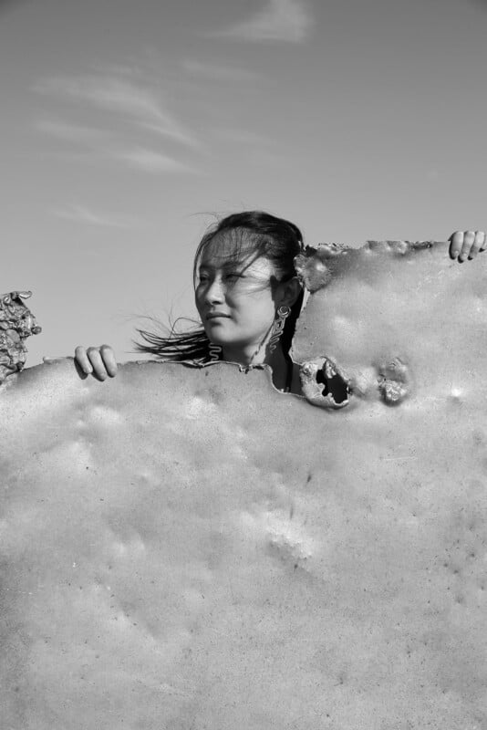 A woman stands behind a large, irregularly shaped rock or sheet with holes, gazing to the side; her hair is blowing in the wind. The image is in black and white with a clear sky background.