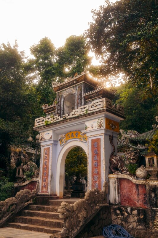 An ornate, traditional stone gate with colorful decorative details stands at the entrance to a temple surrounded by lush trees and greenery in soft sunlight. Stairs lead up to the arched doorway.