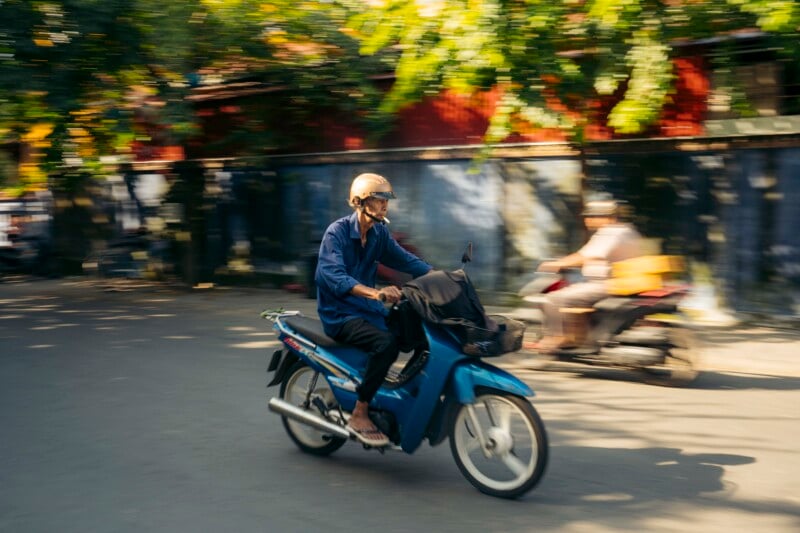 A person wearing a helmet and blue clothing rides a blue scooter on a sunny street, with motion blur in the background and another rider on a scooter passing by.
