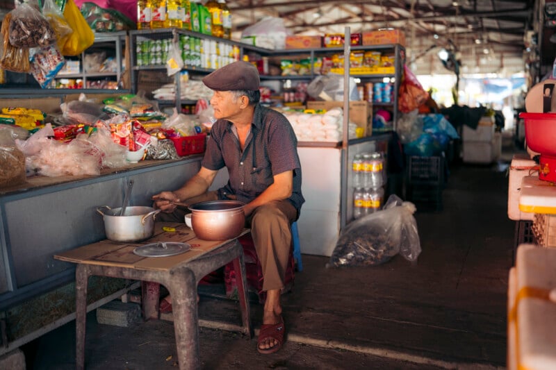 An older man in a flat cap sits at a stall in a market, surrounded by shelves of food and supplies. He holds a spoon over pots on a small table, appearing to prepare or eat a meal. The market is dimly lit and busy.
