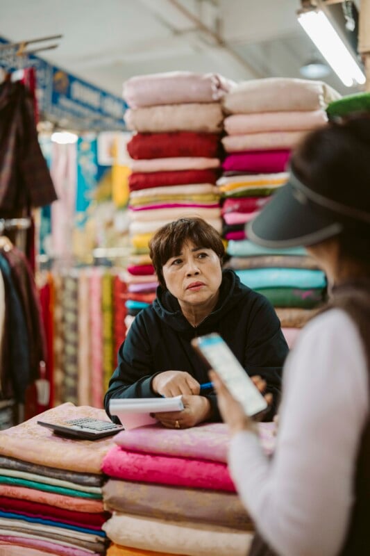A woman sits behind a stall with colorful fabric stacks, holding a notebook and pen, while another person stands in front of her holding a smartphone, possibly discussing or negotiating.