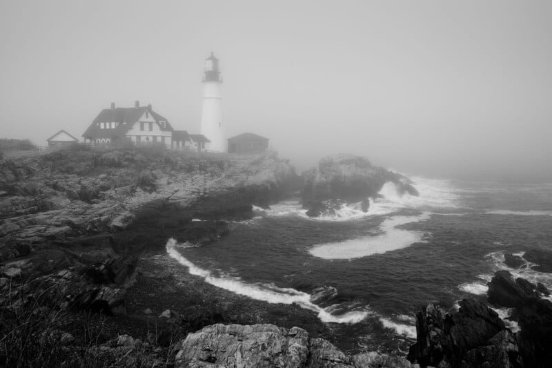 A black-and-white photo of a lighthouse and adjacent buildings on a rocky coastline, surrounded by fog and waves crashing against the rocks.