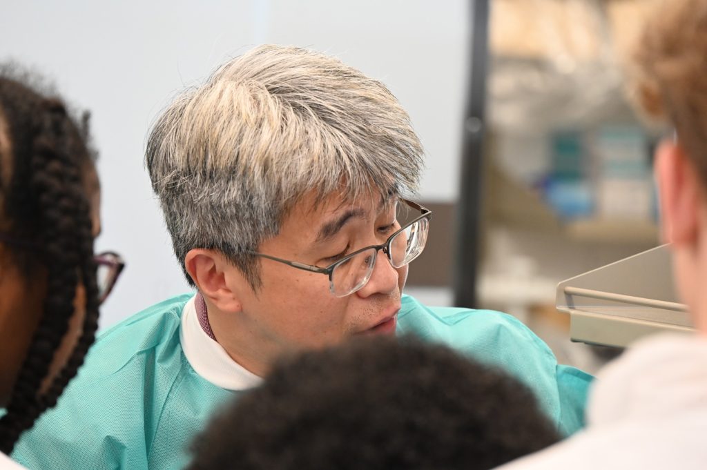 A male scientist in full lab gear speaks with a large group of high school students about the work that happens in his lab.