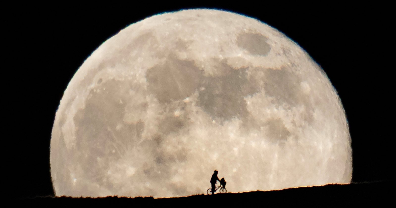 A person on a bicycle with a child silhouetted against a large, bright full moon in the night sky.